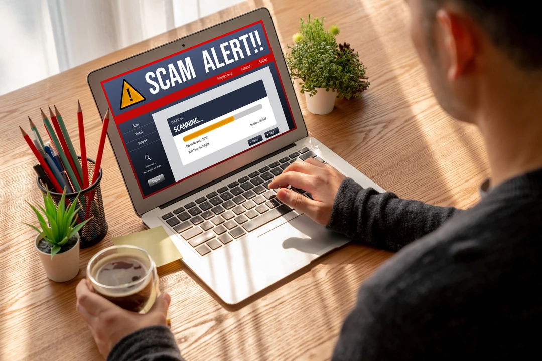 A person sits at a wooden table, viewing a laptop screen displaying a "SCAM ALERT" message while holding a glass of beverage.